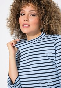 Ljusblå ribbad topp med svarta horisontella ränder. Har en hög halsringning och axeldetaljer, med en avslappnad passform och lösa ärmar.