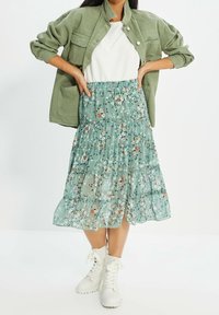 Une jupe superposée arrivant au genou, faite d'un tissu transparent vert avec des motifs floraux blancs, orange et roses. Assortie d'une veste légère et de bottes.