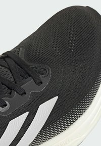 Baskets de sport noires avec une tige en tricot texturé, des rayures blanches et une semelle en maille à motifs. Les lacets sont tissés à travers des œillets renforcés.