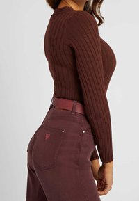 Maglia marrone a maniche lunghe con righe, abbinata a jeans bordeaux a vita alta e una cintura coordinata. Dettagli notevoli includono il tessuto testurizzato e un'amologazione con logo.