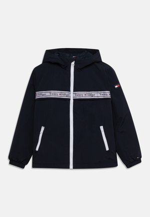 Chaqueta azul marino con capucha, cremallera blanca y bolsillos, puños elásticos y una franja horizontal con el logo de Tommy Hilfiger en el pecho y el brazo.