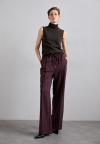 Mouwloze coltrui van breiwerk in donkrustkleur, gecombineerd met losse paarse broek met een elastische tailleband en trekkoord. Puntige zwarte hakken.