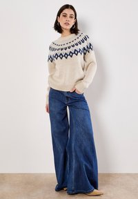 Jersey de punto en color crema con patrones geométricos en azul y negro en el yugo. Combinado con pantalones vaqueros de denim azul de pierna ancha. Corte casual y tejido texturizado.