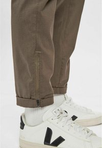 Bruna, texturerade byxor med dragkedjeärmar, parat med vita sneakers med svarta detaljer och snören. Nära vy av fötterna och byxorna.