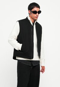 Vesta neagră fără mâneci peste un pulover crem, purtată cu o cămașă în carouri. Accesoriile includ ochelari de soare închiși la culoare. Un outfit casual, stratificat.