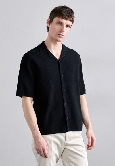 Cárdigan negro de manga corta hecho de tela suave, con cuello muesca y botones en el frente. Llevado sobre pantalones de color claro.