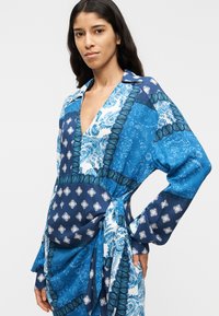 Μπλε φόρεμα με σχέδιο που διαθέτει βαθύ V ντεκολτέ, μακριά μανίκια και ζώνη μέσης με δέσιμο. Το ύφασμα συνδυάζει διάφορες αποχρώσεις του μπλε με διακοσμητικές λεπτομέρειες.