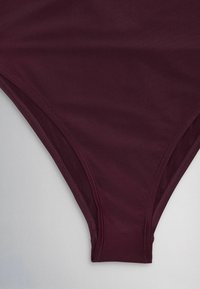 Dark burgundy swimsuit bottom with high-cut leg openings and visible stitching on a light gray background.