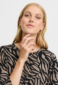 Topp med zebra mönster i tan och svart. Har rund halsringning och trekvartslånga ärmar. Modellen poserar med fingrarna nära hakan och visar upp örhängena.