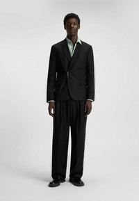 Black tailored suit with a fitted blazer, matching wide-leg trousers, and a light green button-up shirt underneath. Smooth fabric texture.