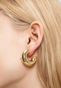 Goldener, geschwungener Creolenohrring, der an einem Ohr präsentiert wird. Er besitzt eine glänzende, glatte Oberfläche mit einem kräftigen, markanten Design und einem kleinen zusätzlichen Steckerohrring.