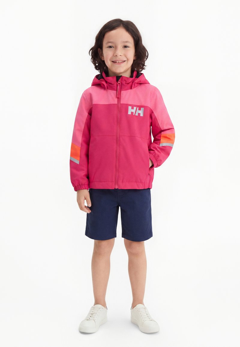 Enfant aux cheveux bouclés et foncés portant une veste à capuche rose et rouge, un short bleu marine et des baskets blanches, souriant et debout avec une main dans la poche.