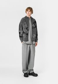 Cardigan grande de color gris oscuro con patrones negros texturizados, camiseta gris y pantalones anchos de gris claro, combinados con zapatos negros slip-on.