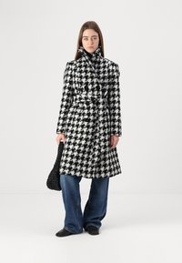 Melns un balts houndstooth raksta mētelis ar jostu viduklī, dubultpogātu priekšu, platām atlokiem un teksturētu audumu. Kombinēts ar džinsa apģērbu un aušanas somu.