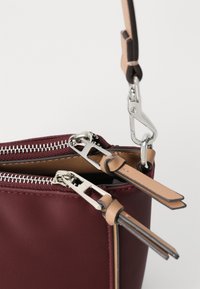 Geantă burgundy cu două fermoare argintii, curele din piele în nuanțe de bej și maro, având o textură netedă. Capsele fermoarului au un design rectangular.
