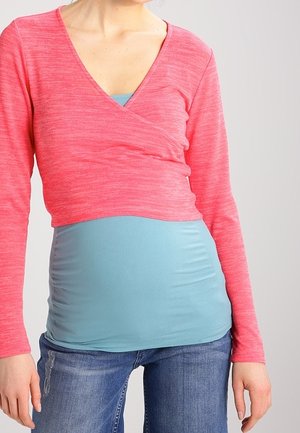 Roze lange mouw wikkeltop met een diepe V-hals over een lichtblauw aansluitend shirt, gecombineerd met blauwe spijkerbroek. Zachte, gestructureerde stof; figuurvolgend ontwerp.