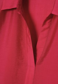 Close-up af en dyb V-hals på et klart rødt tøjstykke lavet af glat, let reflekterende stof med synlige sømme.