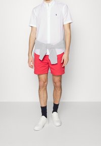 Vit skjorta med krage, röda shorts, grå tröja knuten runt midjan, vita sneakers och mörka strumpor. Enkel, avslappnad klädstil.