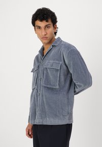 Camisa de pana en gris con un corte relajado, cierre de botón en la parte delantera, dos bolsillos en el pecho y un cuello con muesca. Textura de costillas vertical en toda la prenda.