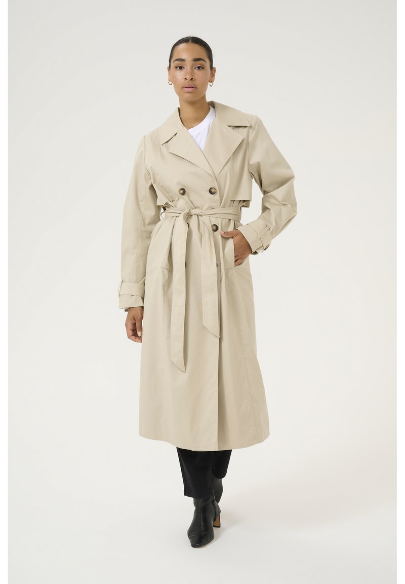 Trench coat color beige chiaro con design doppiopetto, ampio colletto e cintura coordinata. Presenta tasche frontali e lunghezza fino al ginocchio.