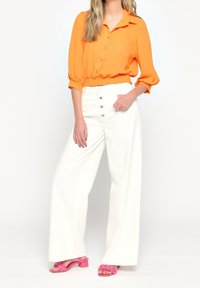Blusa arancione con bottoni e vita elastica, abbinata a pantaloni bianchi a gamba larga con abbottonatura frontale. Indossata con sandali rosa intrecciati.