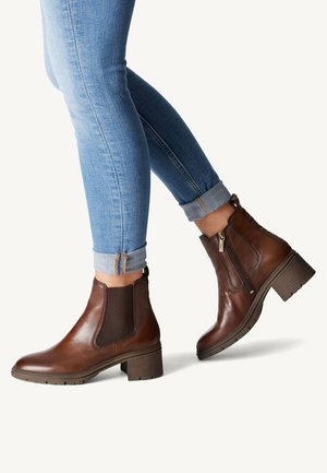Bottines en cuir marron avec un large talon, des panneaux élastiques sur les côtés et une fermeture éclair sur le côté. Portées avec un jean bleu clair, les poignets retroussés.