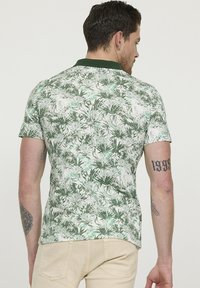 Polo à manches courtes de couleur blanche avec un imprimé botanique vert, doté d'un col vert foncé. Le tissu a une texture lisse et une coupe ajustée.