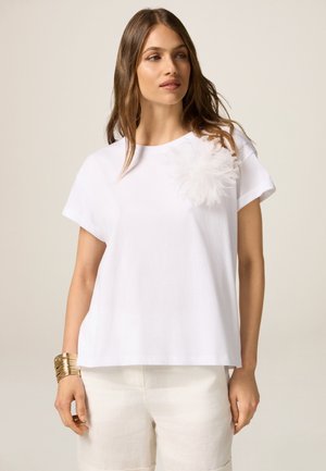 Weißes kurzärmliges T-Shirt mit einem strukturierten weißen Blumenakzent auf der Brust. Lockere Passform, weiches Baumwollmaterial, kombiniert mit hellen Shorts.