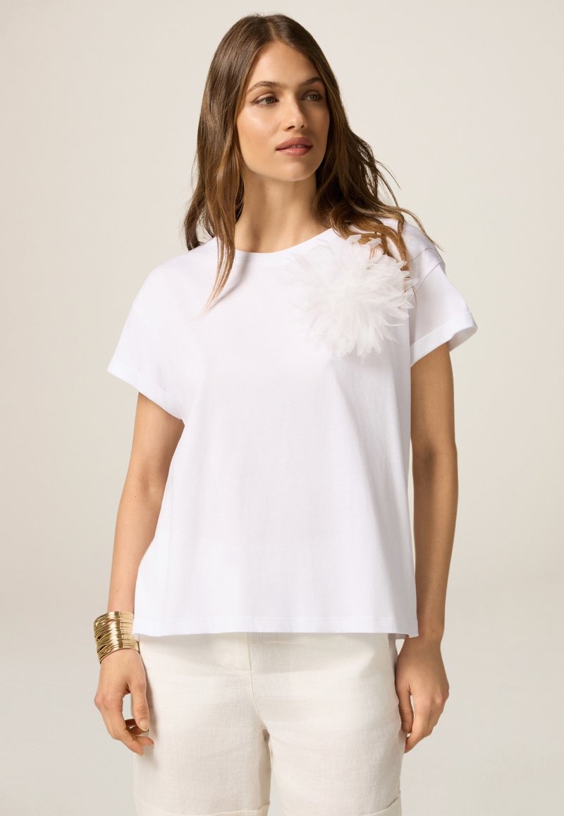 Camiseta blanca de manga corta con un acento floral texturizado en blanco en el pecho. Corte holgado, material de algodón suave, combinada con pantalones cortos de color claro.