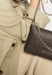 Calvin Kleinin olkalaukku tummanruskeana, kohokuvioidulla logokuviolla, kultasävyisellä ketjuhihnalla ja vetoketjusuljennalla. Kangaspinta on sileä.