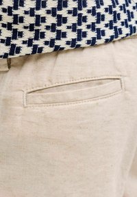 Κοντινό πλάνο από τσέπη μπεζ παντελονιού κάτω από μια μπλε-σκούρα και λευκή υφαντή ζώνη.