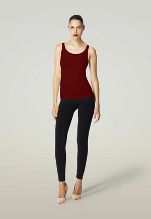 Femme debout portant un haut sans manches rouge foncé, des leggings noirs et des talons hauts beiges sur un fond clair uni.