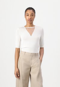 Hvit strikket topp med korte ermer og V-hals, med et gjennomsiktig panel ved halsen. Kombinert med beige høyt liv bukser.