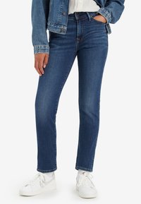 Personne portant un jean slim bleu foncé, une veste en jean, des baskets basses blanches, debout avec une main dans la poche sur fond blanc.