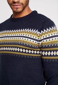 Marinblå stickad tröja med Fair Isle-mönster och vita, gula och grå inslag. Rund halslinning och texturerad tyg.