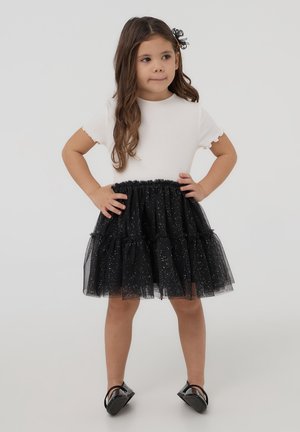 T-shirt bianco a maniche corte con bordi scallop, abbinato a una gonna in tulle nera con dettagli scintillanti e strati di volant. Scarpe flat nere.