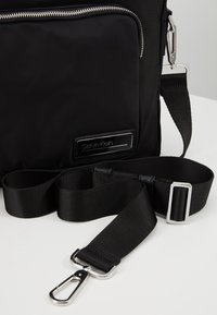 Sac à épaule noir Calvin Klein avec fermeture éclair argentée, sangle noire réglable et boucle métallique sur une surface claire.