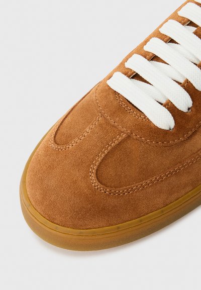 Sneaker in camoscio marrone con lacci bianchi, punta arrotondata e suola in gomma. Presenta cuciture sulla tomaia e una texture liscia.
