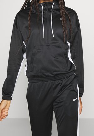 Person iført sort og hvid Reebok tracksuit med halvlange lynlås-hoodie og bukser med elastik i livet, stående mod en ensfarvet baggrund.