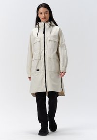 Cappotto lungo beige chiaro con zip anteriore, quattro grandi tasche e cappuccio. Presenta cordini e dettagli neri, abbinato a pantaloni neri.