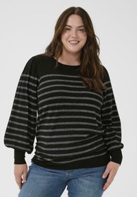 Mujer sonriente con pelo largo castaño, vistiendo un suéter a rayas negro y gris y unos vaqueros azules, de pie frente a un fondo liso.
