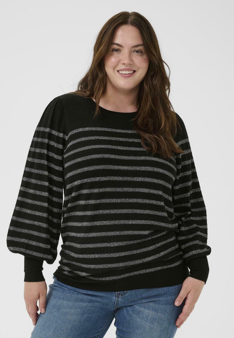 Mujer sonriente con pelo largo castaño, vistiendo un suéter a rayas negro y gris y unos vaqueros azules, de pie frente a un fondo liso.