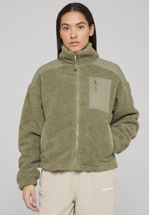 Žena nosi zelenu sherpa flis jaknu s visokim ovratnikom, prednjim patentnim zatvaračem i džepom na prsima s patentnim zatvaračem, stoji pred jednobojnom pozadinom.