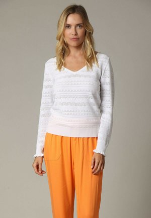 Femme aux cheveux blonds porte un pull blanc à motifs à col en V et manches longues, et un pantalon orange ample, debout devant un fond uni.