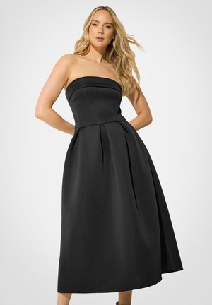 Mujer con cabello rubio largo que lleva un vestido midi negro sin tirantes con falda plisada, de pie frente a un fondo claro y liso.