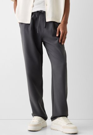 Homme portant un pantalon ample gris foncé à cordon, des baskets blanches, un débardeur blanc et une chemise beige clair à manches courtes, debout devant un fond uni.