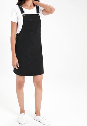 Vrouw draagt een zwarte denim salopetjurk over een wit T-shirt en witte sneakers, staand met één hand die haar nek raakt.