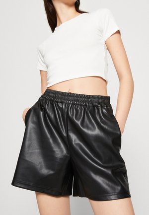 Kvinde iført højtaljede sorte shorts i kunstlæder med elastisk talje og hvid kortærmet cropped top mod ensfarvet baggrund.