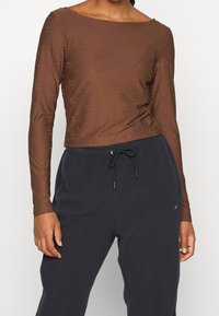 Osoba v těsném hnědém dlouhém rukávu s jemnými logy a černých fleecových kalhotách s tkaničkou v pase a bočními kapsami.