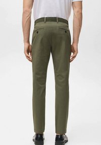 Mężczyzna w oliwkowozielonych spodniach i czarnych butach, stojący tyłem, z rękami swobodnie opuszczonymi wzdłuż ciała, ubrany w białą koszulę z krótkim rękawem.